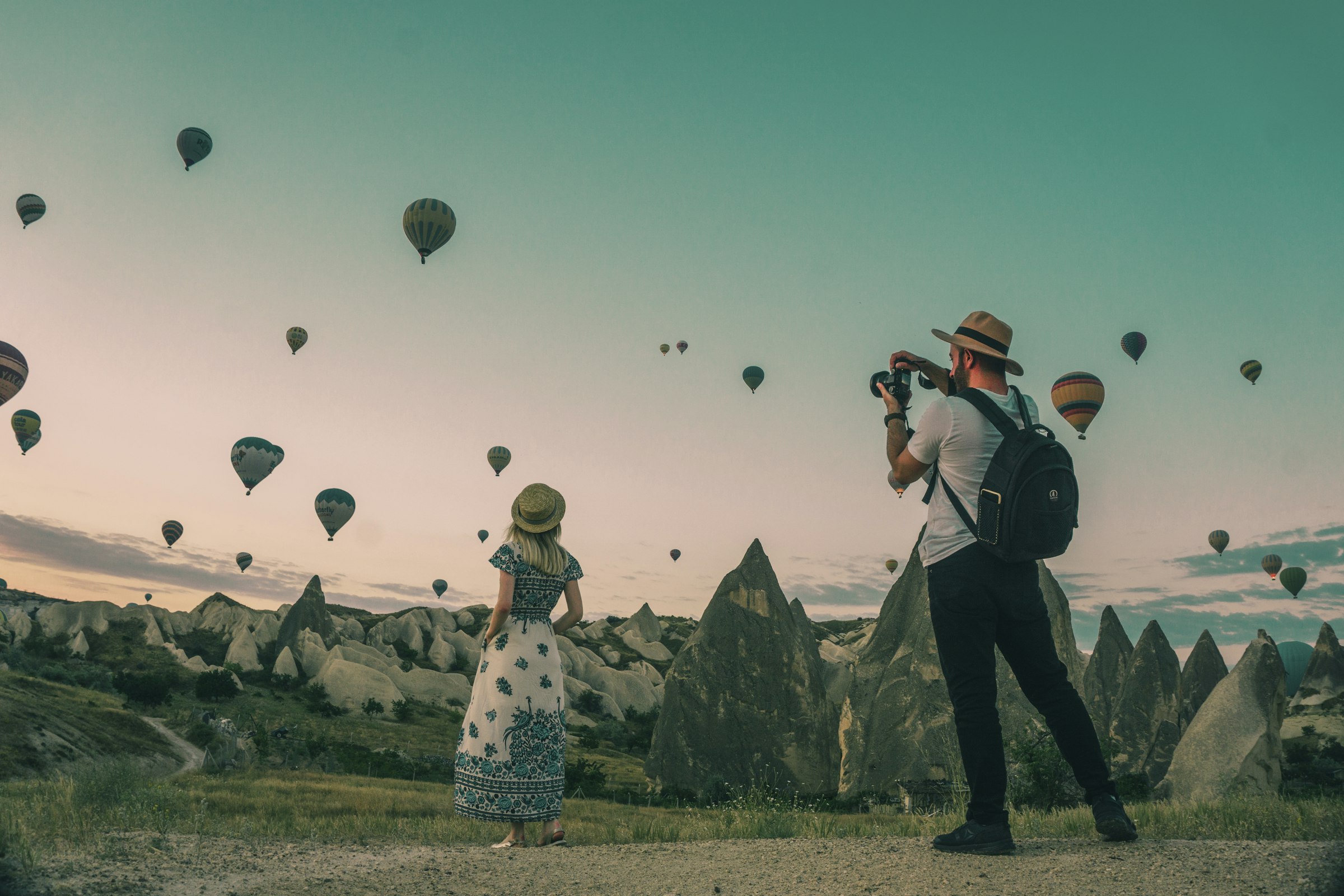 Two travellers beneath a sky filled with hot air balloons at sunrise